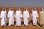 Placeholder: Four Arab sheikhs sitting in the desert wearing typical Arab dress, looking towards the four cardinal points. A talll fat european man in business suit wathhing them while thinking.