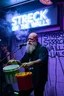 Placeholder: An "Mischief man" performing at dance club, his name is "Janne" bald with beard and looks crazy, Paint buckets on stage, Neon lights and smog, "STRECK ON THE WALLS" SIGN ON WALL, white walls with black paint lines