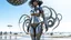 Placeholder: A woman with dark hair in a silver robotic catsuit, standing on a beach with flying mushrooms looking like parasols, with octopus tentacles in the air