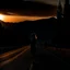 Placeholder: dark night, a woman silhouette on the side of the road,, mountains and forests next to the road, a beautiful orange sunrise in the distance, photo quality