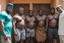 Placeholder: group of big stocky black muscular men from Niger , tank top, bulging shorts, inside a modest living room, queuing in front of the door of a room