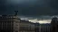 Placeholder: Dramatic, moody cityscape photograph featuring a row of tall, historic buildings under a dark, stormy sky. The layout is vertical, with the buildings aligned in a perspective that leads the eye towards the horizon. The architecture is characterized by a mix of classical and early 20th-century styles, with ornate facades and intricate details. A prominent feature is a statue of an angel atop one of the buildings, illuminated by a beam of sunlight breaking through the clouds. The buildings are pre