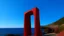 Placeholder: Una prominente estructura roja en forma de T, posiblemente simbólica o artística, se alza audazmente sobre un sereno telón de fondo costero. La figura monolítica tiene una rica textura, evocando un sentimiento de crudeza, materialización orgánica, y contrasta notablemente con la apagada, primer plano terrenal. Este primer plano se funde en las profundidades, extensión ultramar del océano, , que es plácido bajo un cielo suavemente iluminado. Los sutiles toques de luz ambiental crean un efecto res