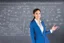 Placeholder: A pretty brown-haired, light-skinned woman in an elegant blue suit stands in a classroom in front of a blackboard with various charts and diagrams, in the sunlight