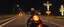 Placeholder: The kebab man driving fast on his motorcycle, fire from the exhaust, widescreen picture, empty road on the country at night, comic style, no helmet and he have a long black beard, neon kebab signs along the road, jesus at his cross