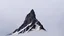 Placeholder: looking up at a single sharp narrow mountain, the peak obscured by clouds and fog.