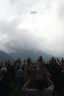 Placeholder: A land with mountain filled with humans everywhere raising hands up for a strands or unexplained object in the sky hovering in a foggy cloudy sky