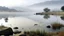 Placeholder: SA wide shot of a lake with a rocky shoreline under a foggy sky. The lake is calm and reflects the sky, with some tall grass growing near the edge. A large pile of rocks forms a barrier or dam along the right side of the frame, sloping upwards. Several people are sitting or standing on the rocks near the water's edge. In the distance, mountains are visible through the fog. Streetlights line the top of the rocky barrier, disappearing into the fog. The color palette is muted, with shades of gray,