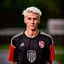 Placeholder: 85mm DSLR color photography of a very detailed headshot fitting all of head and hair in frame. 18-year-old Austrian soccer player, and with no facial hair and has no facial hair, has very short length straight white hair with a small smile, grey background,