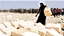 Placeholder: A Palestinian woman wearing a dress carrying very large bags of flour on her back, bending her back down in the destroyed Gaza City, and aid boxes descending from planes near the sea, with a large number of children looking up.