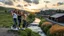 Placeholder: a group of young ladies in sports pants and blouse are dancing to camera in village over high grassy hills,a small fall and river and wild flowers at river sides, trees houses ,next to Ripe wheat ready for harvest farm,windmill ,a pretty train is arriving to station,cloudy sun set sky