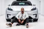 Placeholder: The image shows a man sitting on the floor in front of a futuristic-looking silver Tesla Cybertruck. The man is wearing a white jacket, black pants, and black shoes, accessorized with a chain necklace. The setting appears to be a sleek, minimalistic indoor showroom with white floors and walls, giving the scene a modern, high-tech atmosphere.