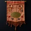 Placeholder: an autumn colored textured cloth hanging with embroidered ornamental leaves and cows, small blank oval brass engraving plate in upper middle, banner is downward pointed bottom, on dark background, western style