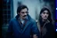 Placeholder: a young man and a beautiful woman standing next to each other, 1 9 8 0 s analog video, with mustache, small glasses, cold scene, out of focus background, house on background, the woman has long dark hair, photo realistic