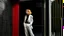 Placeholder: A medium shot captures a modern architectural scene, focusing on a red-framed window that offers a glimpse into an interior space where a Caucasian woman, around 35 years old, stands. She has fair skin, blonde hair, and is dressed in a white jacket and dark pants, complemented by black heels. She is positioned near a wall, seemingly looking out of another window or doorway. The interior features white walls and a dark floor, with a curved wooden structure adding a touch of modern design. The ext