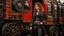 Placeholder: full-length portrait of a pale-faced steampunk woman with auburn wavy shoulder-length hair, with detailed metal arms and legs, dressed in leather trousers, and a jacket, standing beside a steam carriage