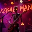 Placeholder: a very thin swedish man (near skeleton), (fat glowing eyes), he is called "KEBAB MAN" performing at death metal club, his name is "KEBAB MAN" kebab rolls On stage, kebab Neon lights and smog, and "KEBAB MAN" big sign on the wall, kebab tribal tattooes, spinning kebab machines, flames and smoke, chaos, he using gasolin and pour it on the guitar on the stage