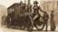 Placeholder: full-height portrait of a pale-faced steampunk woman with straight dark shoulder-length hair, with metal arms and legs, dressed in leather trousers, and a jacket, in a busy Victorian street next to a steam car