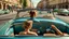 Placeholder: A woman in the foreground seen from the back seat is sitting behind the wheel of a vintage convertible car, looking through the windshield at a busy street scene of an old European city with people and buildings in the background