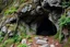 Placeholder: Representación de la entrada a las grutas en la ladera de una montaña, semioculta por vegetación y enredaderas. bosque encantado, magic y fantasia medieval. misticismo sagrado. atmosfera mistica. Formaciones rocosas naturales enmarcan la entrada. La imagen sugiere misterio y aventura, con un contraste entre la oscuridad de la cueva y los colores vivos del exterior. Las Grutas de Anwyn se adentran en las profundidades de la montaña, con entradas ocultas entre vegetación y formaciones rocosas imp