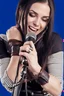 Placeholder: attractive female country music singer leaning forward while holding microphone in front of them, change clothing to plaid in natural tones, leather bracelets on wrists, long hair, mouth open, singing, rings on fingers