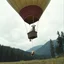 Placeholder: Silent era Art film aesthetic, Long shot Wide angle photograph from perspective of the ground, an excited sheep standing in the basket of a hot air balloon high in air with a scared pig hanging from a rope dangling down from the basket, forest and mountains in the background, cloudy skies, color photo, low contrast, absurd, lots of empty space