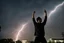 Placeholder: A young man standing, with arms raised, in front of a building at night, with red fork lightning