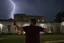 Placeholder: A young man standing, with arms raised, in front of a building at night, with RED fork lightning