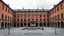 Placeholder: The desolate historic center of a gray city at midday, cloudy and devoid of cars and pedestrians. A civic palace of exposed brick and stone. Thick walls and deep, symmetrical window openings. Axial order and a hierarchy of interior spaces visible from the outside. Expressiveness provided by the rhythm of the openings and the tactile austerity. Contrast between a massive exterior and an introspective void in the central square courtyard. Graphic style: an architectural watercolor by Thomas Schall