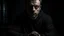 Placeholder: In the photo, we see a man sitting in the dark, looking at the camera with a cold gaze. The man is dressed in black and seems uninterested in what is going on around him.