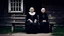 Placeholder: gloomy-looking old women sitting in black hungarian villager dress and wearing east european black head scharf on wooden bench in front of white old house outside in an authentic east european ,hungarian village, high detalied, professional photo, high qualit, high textures. The high-resolution image captures the essence of authenticity and realism, transporting the viewer to another time and place.