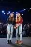 Placeholder: Norwegian Identical teenage twins celebrites, same sex gender standing on a stage with holding their hands, lot of photographers, flashing lights, identical twins with same sex gender