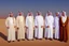 Placeholder: Four Arab sheikhs sitting in the desert wearing typical Arab dress, looking towards the four cardinal points. A talll fat european man in business suit wathhing them while thinking.