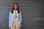 Placeholder: A pretty brown-haired, light-skinned woman in an elegant blue suit stands in a classroom in front of a blackboard with various charts and diagrams, in the sunlight