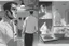 Placeholder: black and white storyboard, wide, on the Foreground there is a man in profile close to the camera and in the background, 3 chefs, scattered throughout the kitchen cooking, frying, cutting