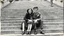 Placeholder: retro monochrome photo of a young hungarian couple in 60s style outfit sitting on the stone stairs in Budapest , the photo is old, stained and scratched in some places, one corner of the photo is missing, the girl's and the boy's faces not clearly visible, blured, behind them are many stairs, summer, moody photo