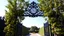 Placeholder: The image is of an ornate gate with a large clock hanging above it. The gate is made of black metal and has intricate designs and patterns on it. On either side of the gate, there are tall trees with green leaves and yellow fruits hanging from their branches. The trees are arranged in a symmetrical pattern, creating a sense of balance and harmony. In the background, there is a pathway leading to a garden with a tree in the distance. The sky is blue and the overall mood of the image is peaceful a