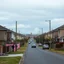 Placeholder: a glimpse of the suburban street, everything is odd, the landscape seems to be made of felt, pastel colours, some people and cars, rather eerie, sinister, void, liminal