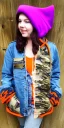 Placeholder: Brunette taking selfie.thick thighs,thick calves,flat belly,curvy fell. big head. Mantle is sewed of upcycled Denim and sewed together of camouflage pieces. Pieces' color are orange, cream and purple. It is with big bright purple felt tippet and redochre-colored-hood is merged with colorful beanie. Big colored headphones (gold rings!) is merged with small felt cap with small visor. Style: Haute Couture in 1950's Africa, N.Y.C fashion in 2023