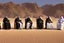 Placeholder: Four Arab sheikhs sitting in the desert wearing typical Arab dress, looking towards the four cardinal points. A talll fat european man in business suit wathhing them while thinking.