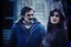 Placeholder: a young man and a beautiful woman standing next to each other, 1 9 8 0 s analog video, with mustache, small glasses, cold scene, out of focus background, house on background, the woman has long dark hair, photo realistic