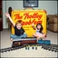 Placeholder: vintage 35mm photograph captures a yellow, rusted metal toy box with red script text reading "THE TROLLEY PROBLEM" with byline in smaller text "WHO LIVES? YOU DECIDE!" placed in a living room. One red-haired boy and one dark haired girl with smiling, exuberant expressions sit cross-legged on a carpeted floor with one arm raised in the air in jubilation. In front of the children, an electric model train lies crashed on its side and smoking near a fork in the train tracks where 5 plastic minifigs