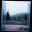 Placeholder: A polaroid photo of an odd room with structures. A transparent tarpaulin covers an image resembling a ruined building. There is a group of fir trees in the background and a high hill in the distance. The surface below is in a pastel color. Fog is present.