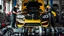 Placeholder: A car repair shop where two people in work clothes are repairing a black and yellow BMW M6 series with an eye-catching theme in a sophisticated and special repair shop where a collection of car engine parts is arranged on the floor at the bottom of the image.