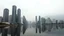Placeholder: A cityscape of tall, modern skyscrapers reflected in a calm body of water, with mountains in the background under a hazy, overcast sky
