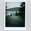 Placeholder: A polaroid photo of an odd room with structures. A transparent tarpaulin covers an image. There is a group of fir trees in the background and a high hill in the distance. A lake. The surface below is in a pastel color. Fog is present.