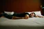 Placeholder: man lying on back on motel bed, arms at sides, face partially turned away, neutral expression, unresponsive posture, plain bedding, generic sheets, harsh flash lighting, dim uneven room lighting, off center framing, candid documentary photo, slightly grainy, no emotion
