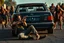 Placeholder: A gritty post-apocalyptic action scene featuring a lone survivor sitting injured on cracked asphalt, leaning against the rear of a bullet-riddled black Mercedes E500 with its back window shattered. The survivor, a rugged young man with long dark hair and a determined expression, aims a handgun toward approaching danger. Surrounding the car are a group of terrifying zombies with decayed skin, tattered clothes, and menacing poses, closing in from all directions. Early morning blue light casts long