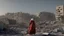 Placeholder: A Palestinian woman wearing a dress carrying very large bags of flour on her back, bending her back down in the destroyed Gaza City, and aid boxes descending from planes near the sea, with a large number of children looking up.