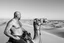 Placeholder: man looking like "Walter White" in breaking bad tv show, bald and goatee, with bare chest riding on a camel in sahara in desert, panorama, black and white photo, staring at a small town in horizon